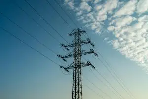 A power line tower against a clear blue sky with scattered clouds, illustrating energy infrastructure.